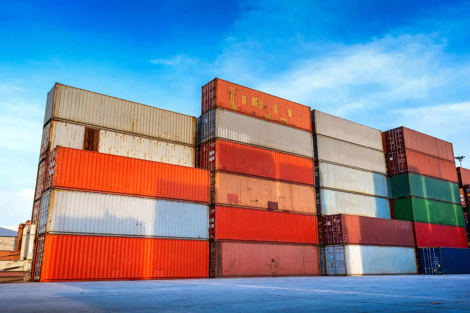 Multiple stacks of colorful shipping containers, including red, orange, white, and green, are piled high in a container yard, representing a large stock of cargo worthy containers ready for use under a bright blue sky.