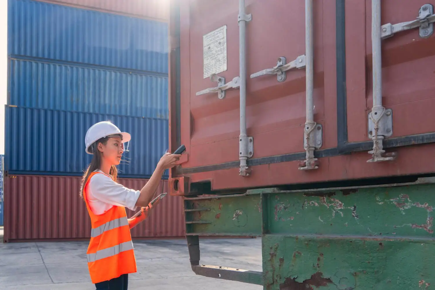 A female logistics worker in a hard hat and high-visibility vest inspects a red shipping container with a scanner and tablet, checking its status as one of many cargo worthy containers at a depot.