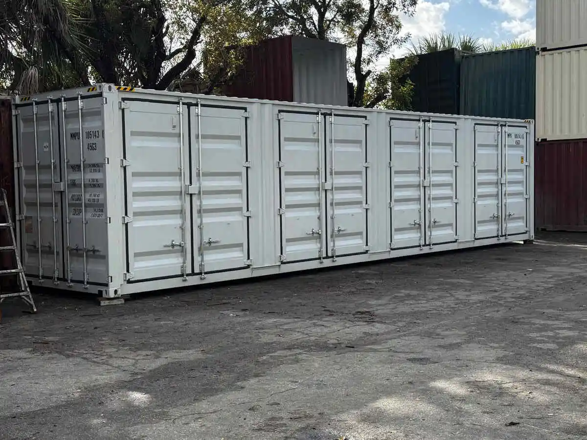 A long, white, open-side shipping container, featuring multiple sets of double doors running along the entire length of one side. The container is sitting on the ground in a storage yard with other colored shipping containers visible in the background.