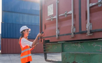 A female logistics worker in a hard hat and high-visibility vest inspects a red shipping container with a scanner and tablet, checking its status as one of many cargo worthy containers at a depot.