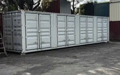 A long, white, open-side shipping container, featuring multiple sets of double doors running along the entire length of one side. The container is sitting on the ground in a storage yard with other colored shipping containers visible in the background.